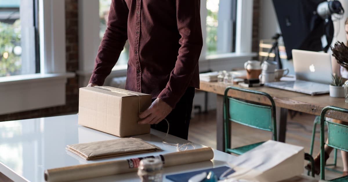 Person packt an einem Tisch ein Paket für den Versand, typische Small-Business-Szene eines Online-Shops